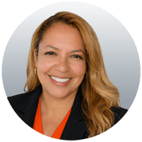 Jeannie is a happy dark skinned woman with light colored shoulder-length hair and is wearing an orange blouse with a black blazer.