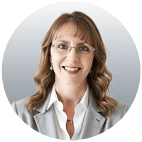 Carol is a happy woman wearing a white collard shirt with a light grey blazer, has light brown curly hair, and is wearing glasses. Carol is a happy woman wearing a white collard shirt with a light grey blazer, has light brown curly hair, and is wearing glasses.