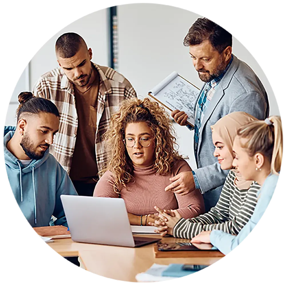 A college classroom of diverse students with teacher surrounding a laptop and discussing study material. A college classroom of diverse students with teacher surrounding a laptop and discussing study material.
